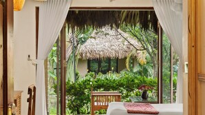 A room with a bed, a wooden chair, a table, and a view of lush greenery and a thatched roof structure. - Cuc Phuong Mineral Retreat (Cuc Phuong)