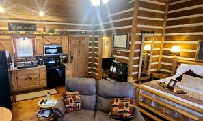 A cozy cabin interior with a kitchenette, a sofa with patterned cushions, a bed with a white comforter, and wooden walls. - Peaceful Mountain Cabin with Mt Yonah Views with Fire Pit and Outdoor Dining in Sautee Nacoochee, Georgia (Sautee Nacoochee)