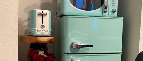 A teal vintage refrigerator with a microwave on top, a wooden shelf with a vase and red flowers, and a small toaster.
