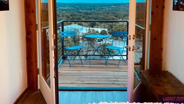 A room with a wooden door leading to a balcony with a table and chairs, a view of the landscape, and a white fluffy rug. - Unique Container Escape with Panoramic Hilltop Views in Cottonwood Shores, Texas (Cottonwood Shores)
