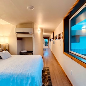 A hotel room with a bed, a window with a view, a hallway, and a door leading to another room.