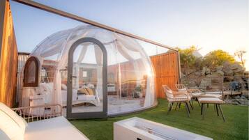 A transparent dome structure with a bed and seating area inside, surrounded by a wooden fence and outdoor furniture.
