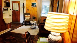 A cozy room with a wooden floor, a green table with a lamp, a chair, a small round table, and a door leading to another room. - Charming Lakeside Cabin Rental near Camp Nine Pines, Wisconsin (Ashland)