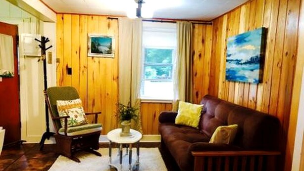 A room with wooden walls, a brown leather sofa, a green armchair, a small round table, and a window with curtains. - Charming Lakeside Cabin Rental near Camp Nine Pines, Wisconsin (Ashland)
