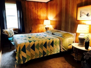 Ein Zimmer mit einem Bett, zwei Nachttischlampen, einem kleinen Tisch mit Pflanze und einem gerahmten Bild an der Wand.