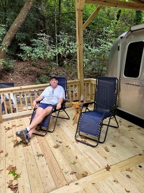 A man in a wheelchair on a wooden deck with a forest backdrop, a small table with a bottle, and a camper parked beside. - Private Wooded Airstream Retreat with Babbling Brook near Cashiers & Highlands – Sapphire, NC (Sapphire)