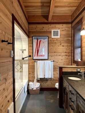 Une salle de bain aux murs lambrissés, équipée d’une douche, d’un lavabo et d’une affiche encadrée.