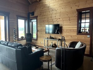 Ein Zimmer mit Holzwänden, einer Ledersofa, einem Fernseher, einem Kamin und Blick durch die Fenster.