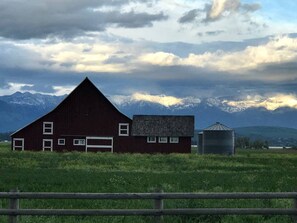 A red barn with a smaller attached building and a silo, set against a backdrop of mountains and a cloudy sky. - Beautifully Renovated Barn Ideal for Glamping at Flathead Lake, Montana (Somers)