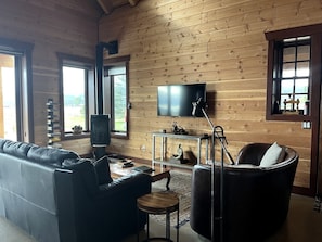 Ein Zimmer mit Holzwänden, einer Ledersofa, einem Fernseher, einem Kamin und Blick durch die Fenster.