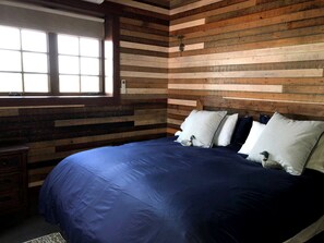 A bedroom with a wooden slat wall, a large bed with blue bedding, and a window with blinds. - Beautifully Renovated Barn Ideal for Glamping at Flathead Lake, Montana (Somers)