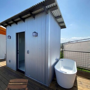 A metal building with a door and two wall-mounted lights, a white bathtub, and a wooden deck. - Glamping Retreat with Outdoor Tub & Panoramic Sky Views in Cottonwood Shores, Texas (Cottonwood Shores)