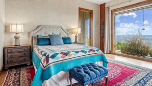 Une chambre avec un grand lit, deux tables de chevet, un banc, une vue à travers une grande fenêtre et une porte donnant sur l’extérieur.