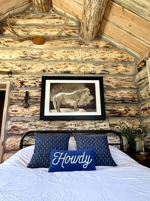 Ein rustikaler Raum mit einer Wand aus Holzbalken, einem gerahmten Bild eines Pferdes, einem Bett mit einem Kissen mit der Aufschrift 'Howdy' und einem Metallbettgestell.