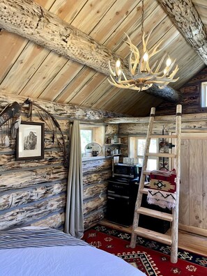 Ein rustikales Cabin-Interieur mit Holzbalkendecke, einer Chandelier, einer Leiter, einem Bett und einem roten Teppich mit Muster.