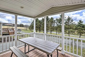 Eine überdachte Veranda mit einem Holztisch und einer Bank, weißen Geländern und Blick auf Bäume und eine Straße.