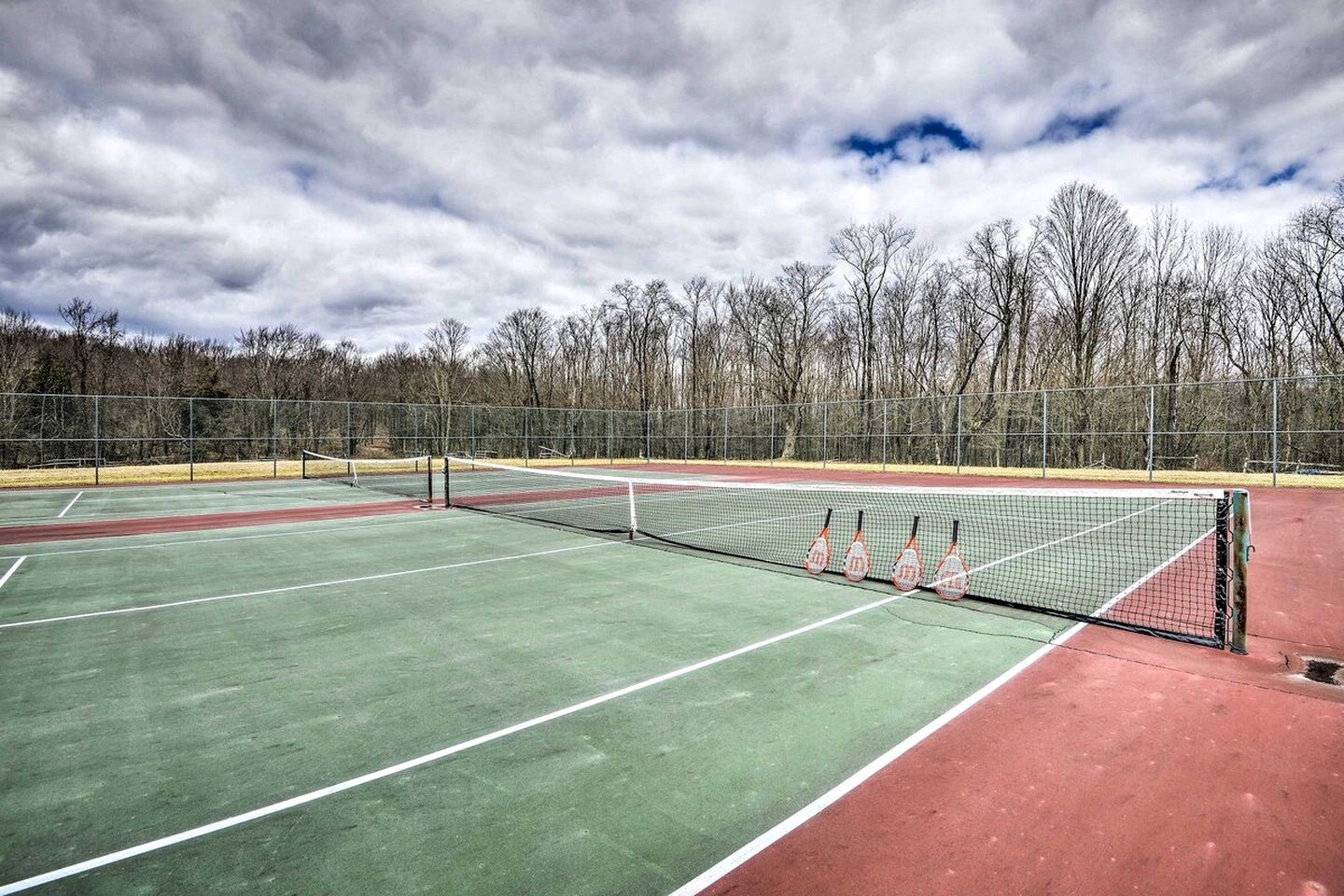 Un court de tennis en plein air, doté d’un filet, entouré d’arbres et surplombé par un ciel nuageux.