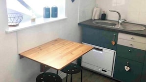Une petite cuisine avec une table en bois et deux tabourets, une fenêtre donnant sur des immeubles et un évier.