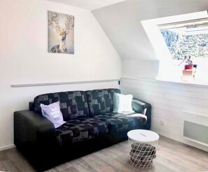 Ein modernes Wohnzimmer mit einem schwarz gemusterten Sofa, einem kleinen runden Tisch, einem Wandbild und einem Fenster mit Blick auf eine verschneite Landschaft.