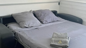 Un lit simple avec une literie et des oreillers gris, une serviette pliée de couleur grise ornée d’un logo, et un fauteuil noir sur la gauche.