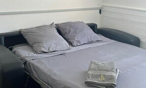 Un lit simple avec une literie et des oreillers gris, une serviette pliée de couleur grise ornée d’un logo, et un fauteuil noir sur la gauche.