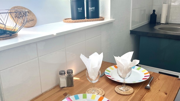 Une cuisine avec une table en bois, deux récipients noirs étiquetés « THÉ » et « CAFÉ », une assiette colorée, un verre avec une serviette, et une vue sur un paysage verdoyant à travers une lucarne.