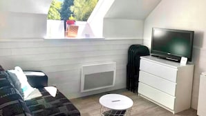 Une petite pièce moderne avec un plafond en pente, un téléviseur encastré, une commode blanche, une table ronde et une fenêtre donnant sur un paysage verdoyant.