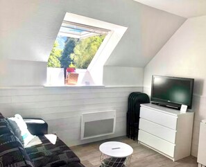 Une petite pièce moderne avec un plafond en pente, un téléviseur encastré, une commode blanche, une table ronde et une fenêtre donnant sur un paysage verdoyant.