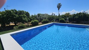 Una piscina rectangular con agua azul cristalina, rodeada de vegetación cuidada y un cielo despejado.