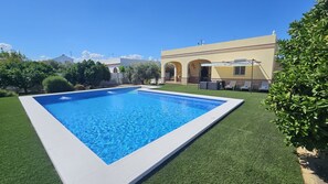 Una piscina rectangular con borde blanco, rodeada de césped artificial verde, y un edificio con fachada amarilla y entrada en arco al fondo.
