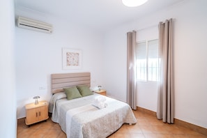 Un dormitorio con cama, mesitas de noche, aire acondicionado instalado en la pared, una ventana con cortinas e imagen enmarcada en la pared.