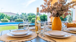 Mesa lista para comer con platos, cubiertos y una botella de vino blanco, un jarrón de flores secas y vistas a una piscina y vegetación.