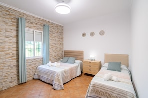 A bedroom with two beds, a stone wall, a window with curtains, a bedside table with a lamp, and a wall clock. - Villa Manantial Country House in the Seville Countryside (La Puebla de Cazalla)