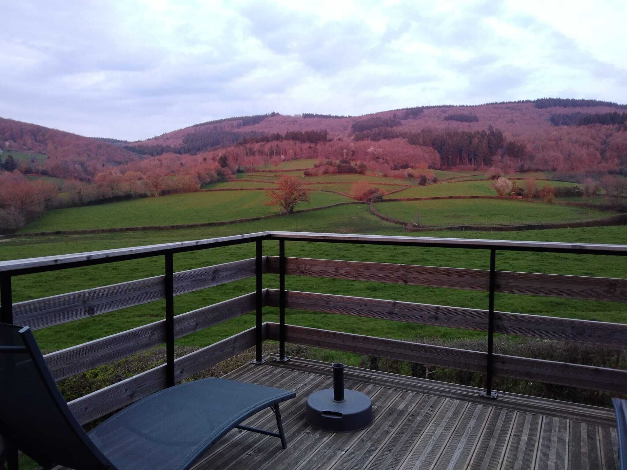 A wooden deck with a metal railing, a single lounge chair, and a view of a hilly landscape with fields and trees.