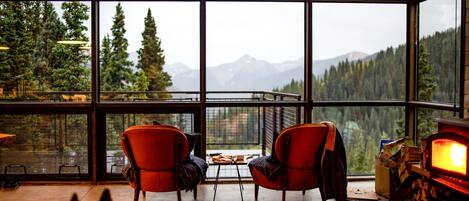 A cozy room with large windows, two red chairs, a table with books and a blanket, and a fireplace.