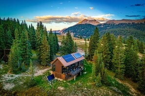 Una cabaña con techo de paneles solares, rodeada de un bosque denso y montañas.