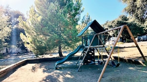 A playground with a slide and swings, surrounded by trees and a clear sky.