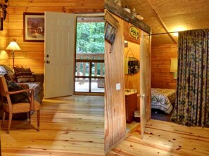 A wooden cabin interior with a bed, a chair, a door leading outside, and a small table. - Cozy Log Cabin for Getaway in the Mountains near Cherokee, North Carolina (Cherokee)