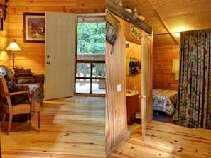 A wooden cabin interior with a bed, a chair, a door leading outside, and a small table. - Cozy Log Cabin for Getaway in the Mountains near Cherokee, North Carolina (Cherokee)