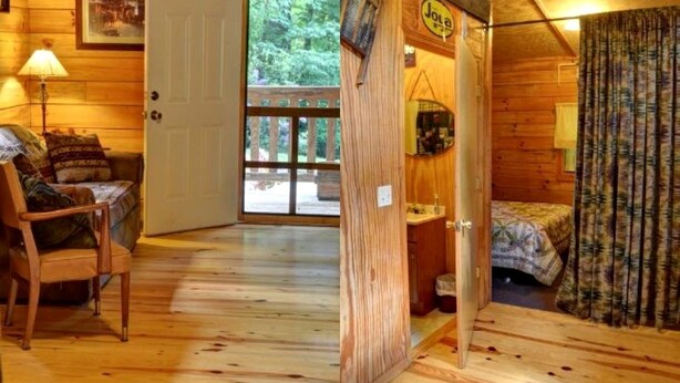 Interior de una cabaña de madera con una cama, una silla, una puerta que da al exterior y una mesita.