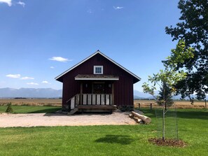 A red barn with a porch, surrounded by a grassy field and mountains in the background. - Luxury Flathead Lake Cabin with Stunning Views (Somers)