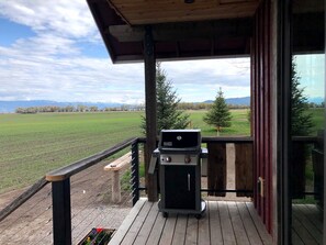 Ein überdachter Außenbereich mit Grill, Holzdeck und Blick auf ein grünes Feld und Berge.