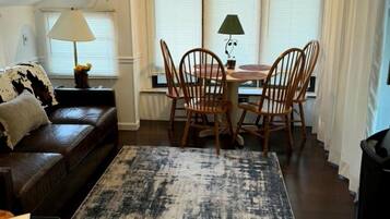 A living room with a brown leather sofa, a dining table with chairs, a patterned rug, and a window with curtains.