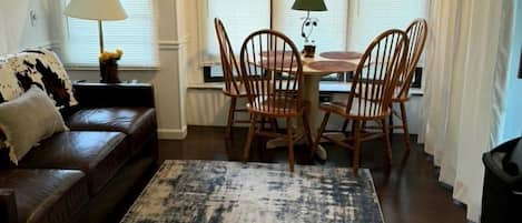 A living room with a brown leather sofa, a dining table with chairs, a patterned rug, and a window with curtains.