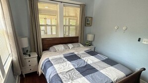 Un dormitorio con cama, mesita de noche, ventilador de techo, ventana con cortinas y un interruptor de luz en la pared.