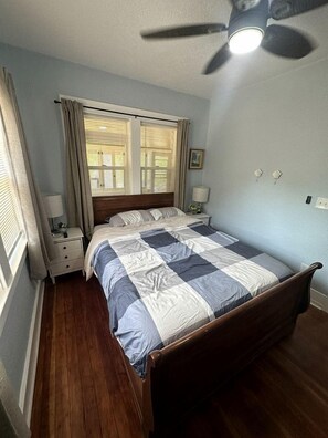 A bedroom with a bed, a nightstand, a ceiling fan, a window with curtains, and a wall-mounted light switch.