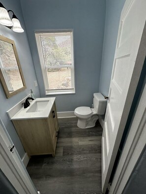 A bathroom with a wooden vanity, a white toilet, and a window with blinds.