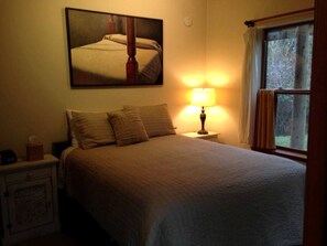 Een slaapkamer met een bed, nachtkastje, lamp, raam met gordijnen en een schilderij aan de muur.