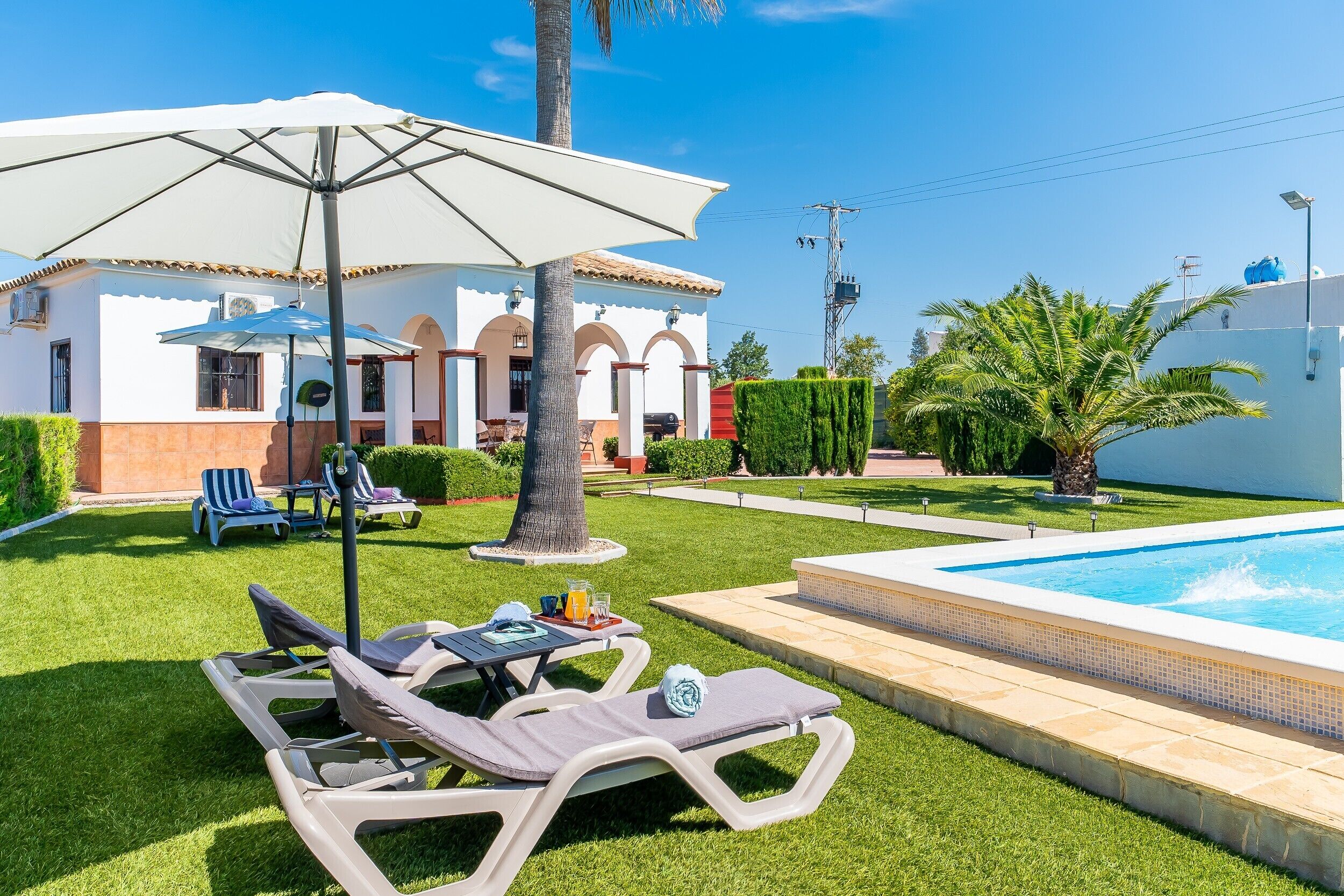 Zona de piscina amb gandules, una taula amb una beguda i un edifici blanc d'arcs.