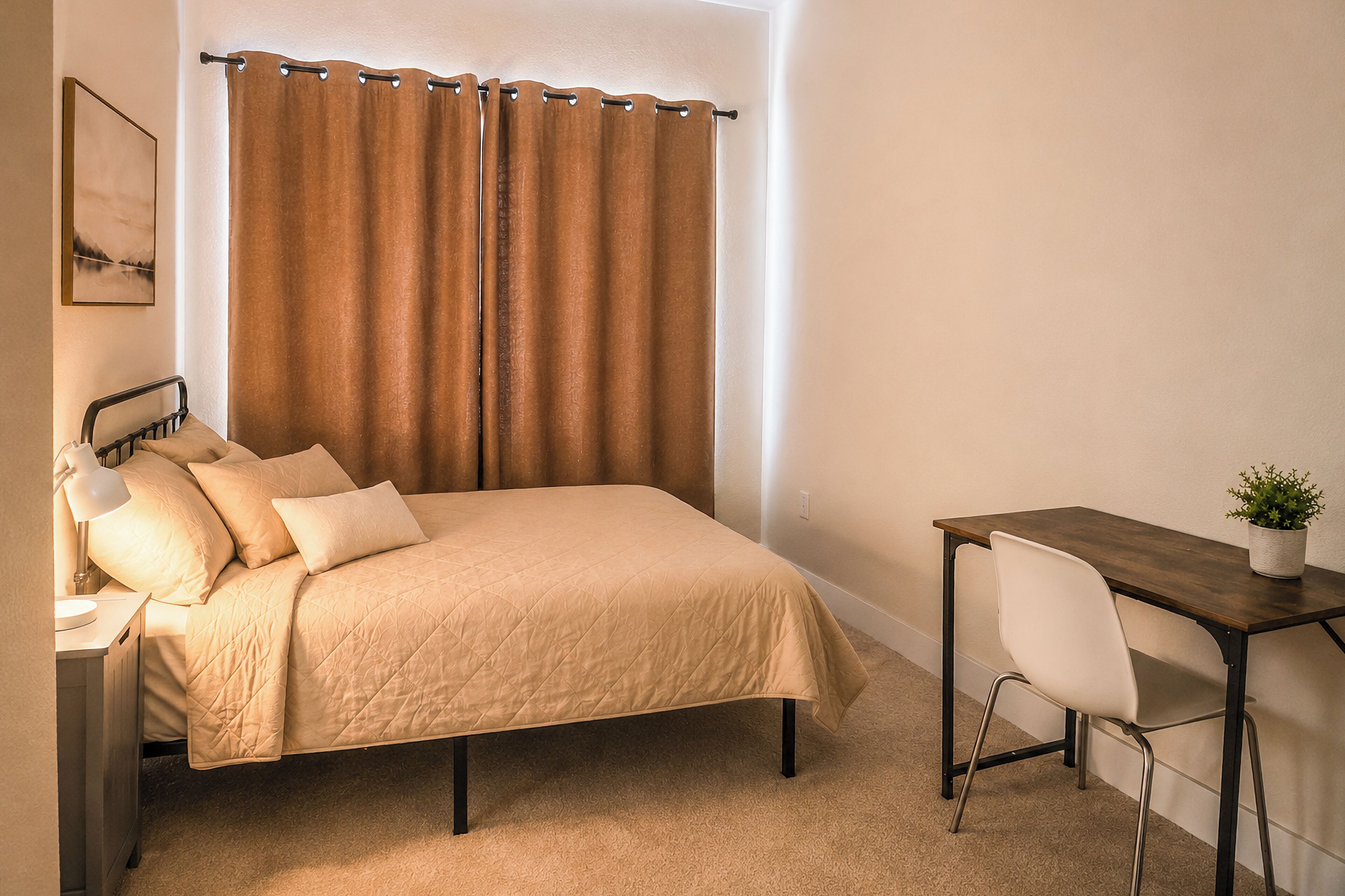 Une chambre à coucher avec un lit, une table de chevet, une lampe et un bureau avec une chaise et une plante.